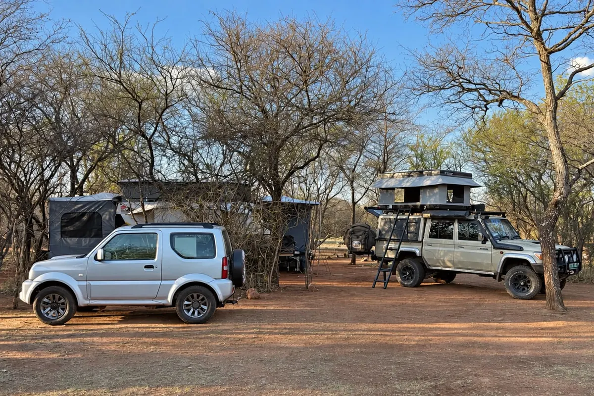 A silver Suzuki Jimny and a Land Cruiser 79 with a rooftop tent, parked on red dirt at a bushveld campsite