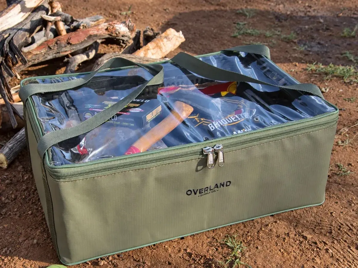 Die Trommel packed with camp kit beside a pile of firewood on red dirt, an axe and briquettes visible through the clear top
